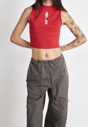 Osoba nosząca czerwony krótki top bez rękawów z napisem „Tommy Jeans” oraz luźne szare spodnie typu cargo, z widocznymi tatuażami na ramionach.