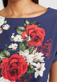 Blusa floreale con uno sfondo blu navy, caratterizzata da rose rosse, gigli bianchi e foglie verdi. Tessuto liscio con scollatura a girocollo e maniche corte.