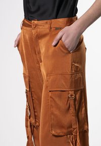 Orange Satin-Cargo-Hose mit Seitentaschen, Knopfverschluss und dekorativen Akzenten. Glatte Textur mit verstellbaren Riemendetails.