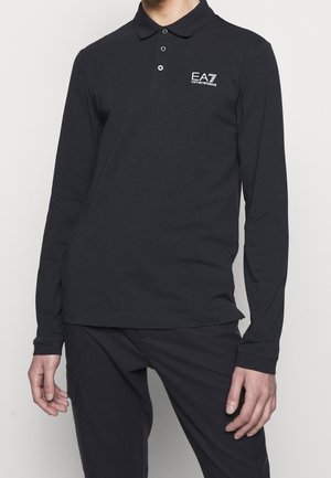 Osoba ubrana w czarną, długorękawową koszulkę polo z logo EA7 Emporio Armani na piersi i czarne spodnie na jednolitym tle.