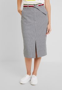 Nainen, joka pukeutuu musta-valkoiseen houndstooth midi-hameeseen, jossa on edessä halkio, raidalliseen vyöhön, valkoisiin tennareihin ja valkoiseen t-paitaan, jossa on keltainen printti.