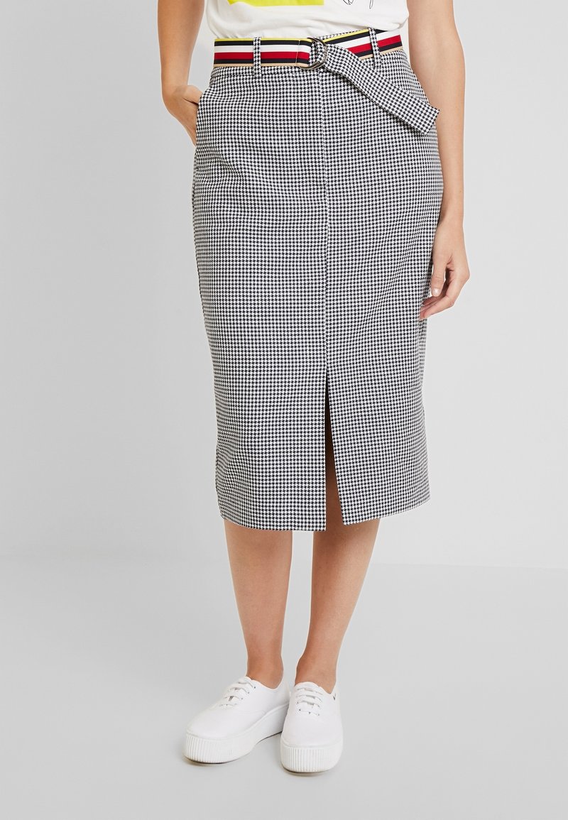 Nainen, joka pukeutuu musta-valkoiseen houndstooth midi-hameeseen, jossa on edessä halkio, raidalliseen vyöhön, valkoisiin tennareihin ja valkoiseen t-paitaan, jossa on keltainen printti.