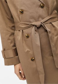 Gabardina beige con botones de carey, cinturón atado en la cintura, tirantes en las muñecas y una mano descansando cerca del bolsillo de la gabardina.
