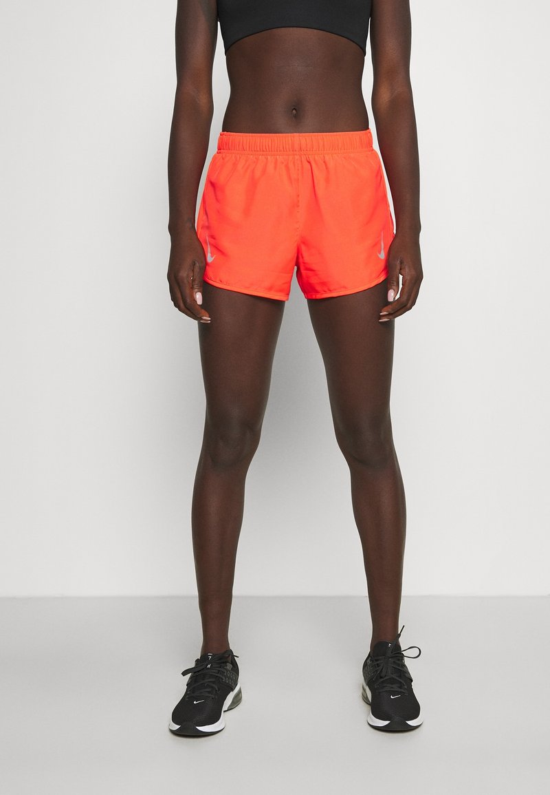 Urheilullinen henkilö, joka pukeutunut kirkkaan oransseihin juoksushortseihin, mustaan treenitoppiin ja mustiin Nike-tennareihin, seisoo yksiväristä taustaa vasten.