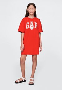 Röd bomullst-shirtklänning med "GAP"-logotyp och hjärtdesign; avslappnad passform, korta ärmar och knälång fåll. Bärs med vita sandaler.