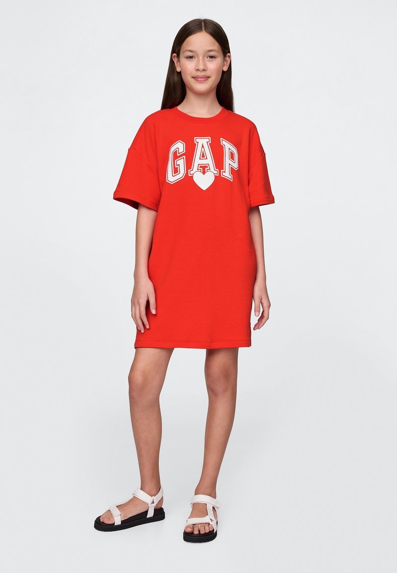 Röd bomullst-shirtklänning med "GAP"-logotyp och hjärtdesign; avslappnad passform, korta ärmar och knälång fåll. Bärs med vita sandaler.