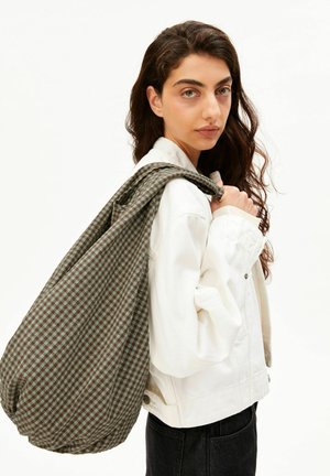 Jonge vrouw met lang donker haar, een wit jack dragend en een grote groenbruine geruite tas over haar schouder, zijwaarts gekeerd.