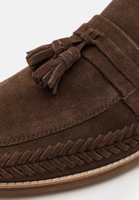 Mocassino in suede marrone con accentuazione a nappina e cuciture decorative intrecciate lungo il bordo, dotato di suola in gomma bianca.