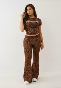 Bruin luipaardprint cropped top en bijpassende flared broek, met een logo op het shirt en een effen tailleband op de broek. Witte sneakers.