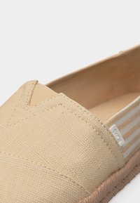 Μπεζ καμβά παπούτσι τύπου slip-on με ελαστικό V-άνοιγμα, ριγωτή υφασμάτινη ταμπελίτσα στη φτέρνα και υφασμένο υφή σόλα.