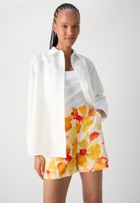 Camicia bianca con bottoni sopra una canotta bianca a coste, abbinata a pantaloni corti a fantasia floreale nei toni dell'arancione e del giallo.