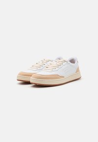 White and beige sneakers with a textured suede overlay, flat laces, and a rubber sole featuring speckles. Subtle branding on the side.