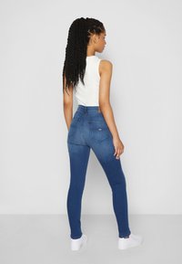 Blue denim skinny jeans with a high waist, featuring two back pockets and a tan leather patch on the waistband. Paired with a white tank top.
