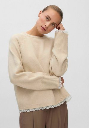 Femme portant un pull beige avec finition en dentelle aux poignets et à l'ourlet, posant la tête inclinée et la main reposant sur la tempe sur un fond uni.
