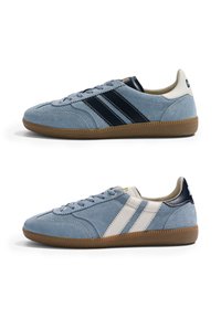 Deux baskets en daim bleu clair avec des semelles en gomme, l'une avec des rayures noires et un soufflet de talon, l'autre avec des rayures blanches et un soufflet de talon.