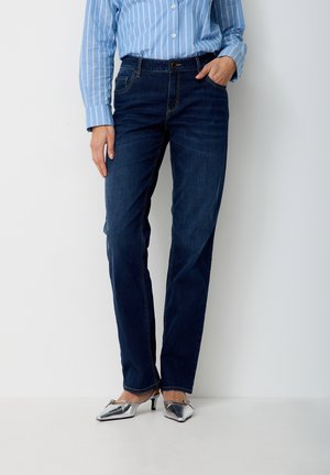 Person trägt dunkelblaue Jeans mit geradem Bein, ein blau-weiß gestreiftes Hemd, das hineingesteckt ist, und silberne, spitz zulaufende High Heels.