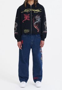 Svart zip-up hoodie med färgglada drake- och ormsgrafik, tillsammans med lösa blå denimjeans med broderade detaljer. Vita sneakers.