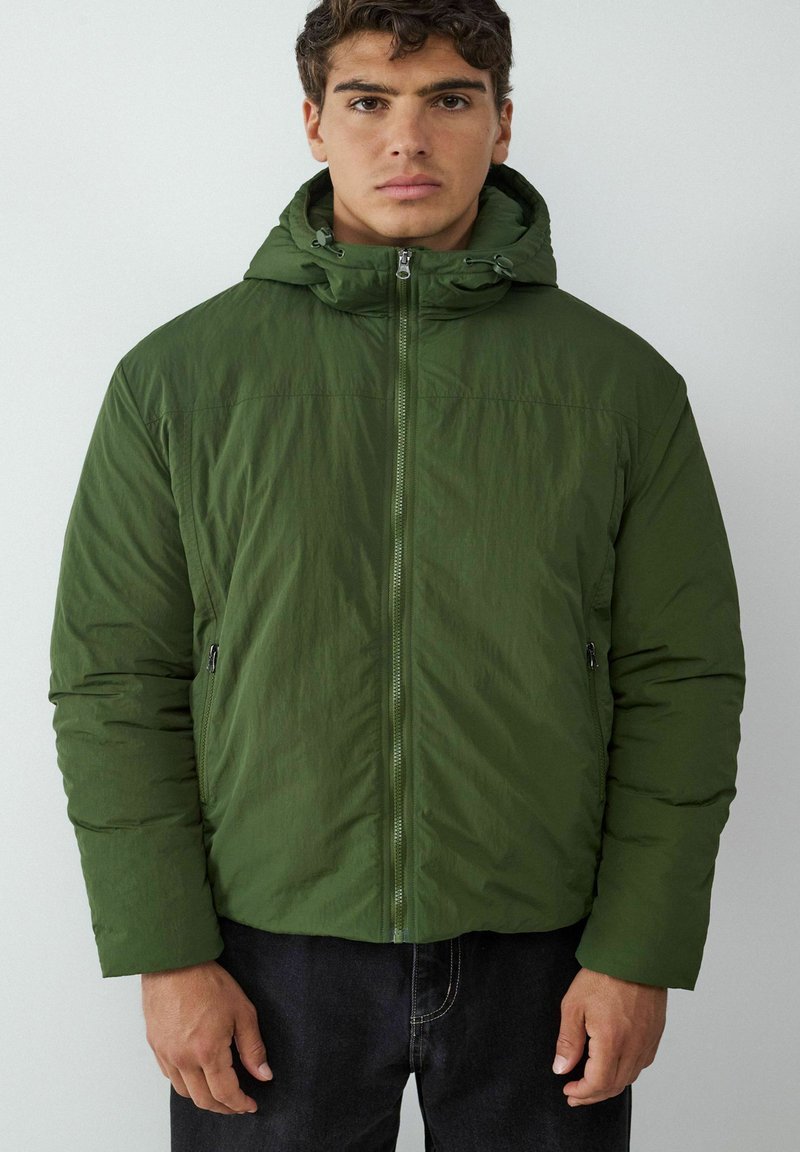 Giacca piumino verde con cappuccio, dotata di chiusura con zip, tasche laterali e superficie texture. Indossata con jeans in denim scuri.