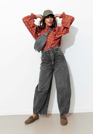 Femme posant avec les mains derrière la tête, portant un chemisier rouge à fleurs, un jean gris à jambes larges, une casquette à imprimé léopard et un sac bandoulière assorti, mocassins marron.