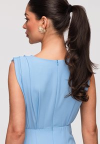 Femme avec une longue queue de cheval noire portant une robe bleu clair sans manches et une boucle d'oreille florale translucide, tournée vers la gauche sur un fond uni.