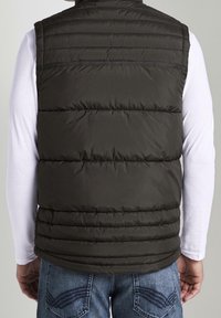 Gepolsterte, ärmellose Weste in dunkelolivgrün mit horizontalem Steppmuster. Tragen über einem weißen Langarmshirt mit hohem Kragen.