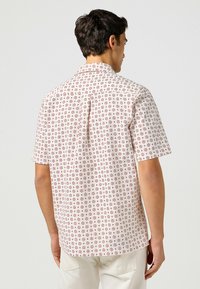 Camicia a maniche corte in bianco sporco con motivo floreale rosso, design con colletto e taglio dritto. Presenta una pettorina posteriore e una texture sottile.