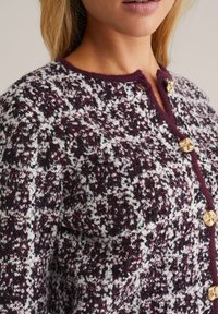 Neulecardigan, jossa on maroon, valkoinen ja musta kuvio, kullansävyiset napitus, pyöreä pääntie ja teksturoitu kangas.