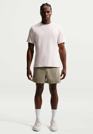 Uomo visto frontalmente che indossa una maglietta rosa chiaro a maniche corte, pantaloncini beige, calzini bianchi e scarpe sportive di colore chiaro su sfondo uniforme.