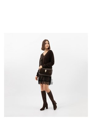 Kobieta ubrana w czarną sukienkę z koronkowym dołem, czarny sweter z dekoltem w serek, brązowe kozaki do kolan oraz ciemnobrązową torbę na ramię.