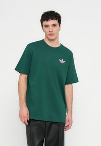 Zaļš kokvilnas t-krekls ar apaļu kakla izgriezumu, īsām piedurknēm un mazu rozā logotipu uz kreisās krūts. Audums šķiet mīksts un gluds.