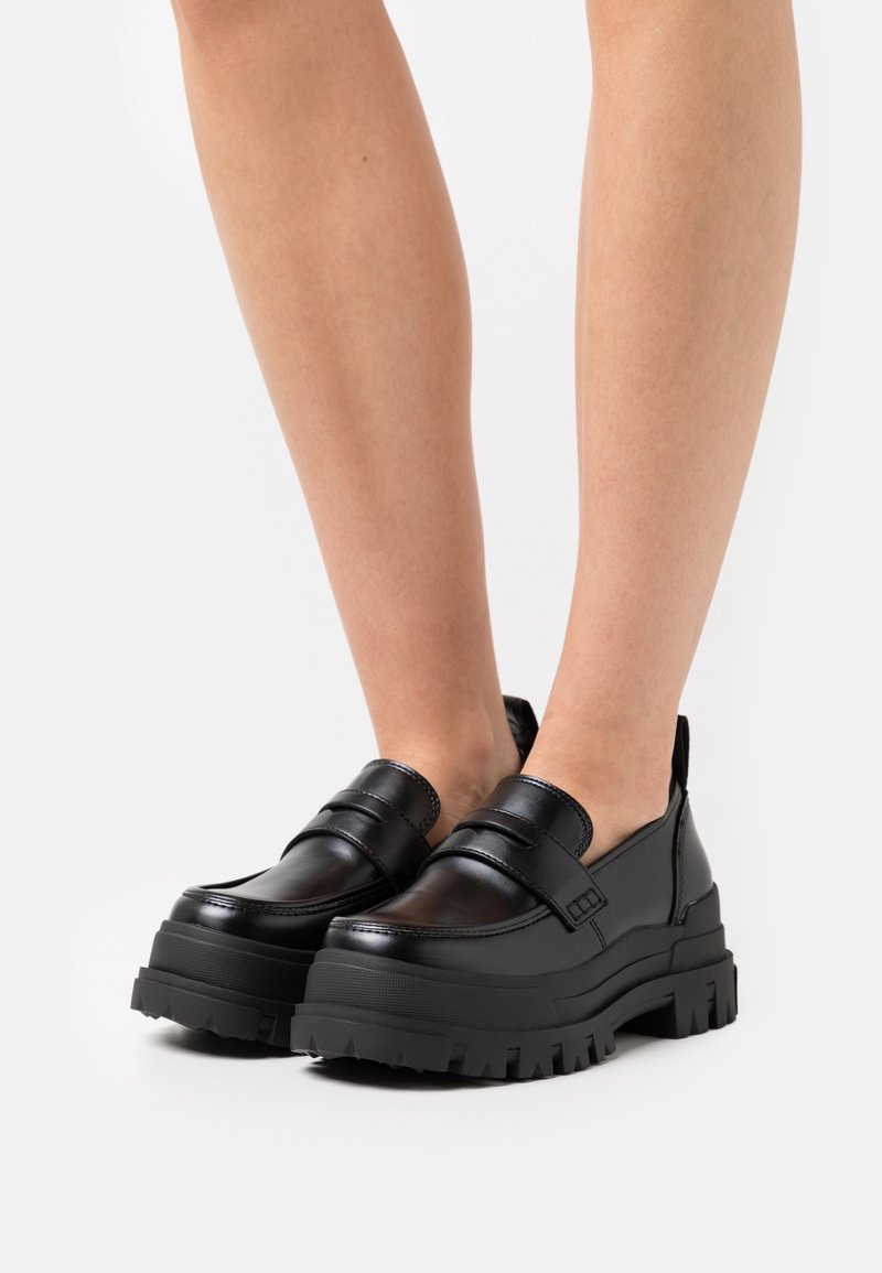 Mocassins à plateforme noire épaisse portés sur des jambes nues sur fond blanc uni.
