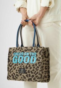 Tote bag im Leopardenmuster mit blauer, glitzernder Schrift "FOCUS ON THE GOOD." Verfügt über zwei blaue Griffe und eine rechteckige Form. Markenlogo sichtbar.