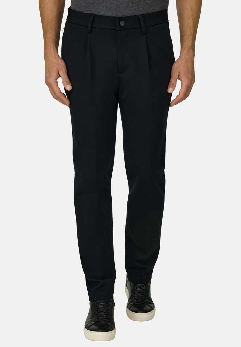 Homme portant un pantalon noir ajusté avec des plis et des baskets noires à semelles blanches, debout devant un fond uni.