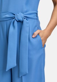Hellblaues Jumpsuit mit geknotetem Taillengürtel, glattem Stoff, weitem Bein-Design und seitlichen Taschen.