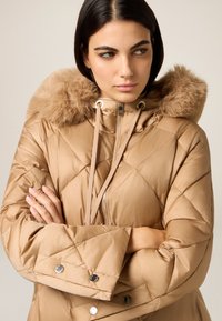 Beige quiltet dunjakke med pelsforet hætte, lynlås, snøre detaljer og metal trykknapper, med en blank finish.