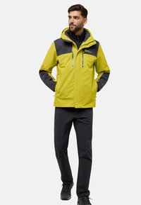 Giacca impermeabile gialla e grigia con cappuccio, tasche con zip e caratteristiche regolabili, abbinata a pantaloni neri e scarponi da trekking.