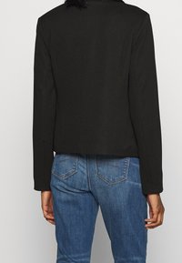 Černý krácený blejzr s dlouhými rukávy, vyrobený z texturované látky a s jednoduchým designem. V kombinaci s modrými džíny.