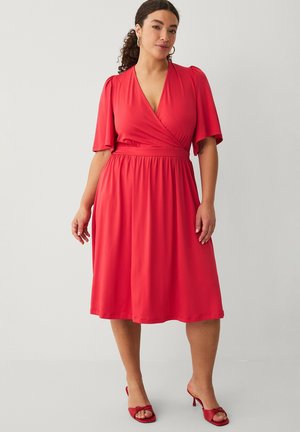 Kobieta z kręconymi włosami w czerwonej sukience kopertowej z falbanowymi rękawami i czerwonych sandałach na obcasie, stojąca na jednolitym tle.