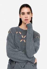 Maglione grigio lavorato a maglia con ritagli floreali, maniche lunghe e polsini a costine. Texture morbida, vestibilità rilassata e aperture significative in tutto il design.