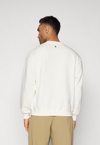 Felpa bianca con colletto rotondo, spalle scese, polsini a coste e un piccolo dettaglio asterisco nero sul retro. Vestibilità casual e texture morbida.