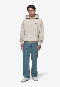 Sweatshirt bege com capuz, bolso frontal e logótipo preto, combinado com jeans azuis claros e sapatilhas brancas com solas castanhas.