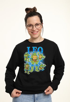 Kvinde iført briller og sort sweatshirt med fem Teenage Mutant Ninja Turtles med teksten "LEO" over dem.