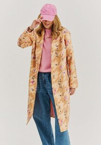 Beige vnější kabát s květinovým vzorem a geometrickým designem; dlouhé rukávy, otevřený přední díl, nošený přes růžový svetr a modré džíny; růžová čepice.