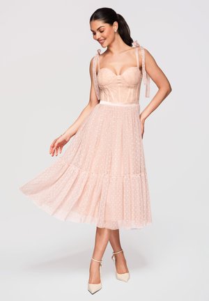 Femme souriante et tournant dans une robe midi en tulle rose clair à pois, avec un corsage ajusté et des bretelles nouées aux épaules, portant des talons blancs.