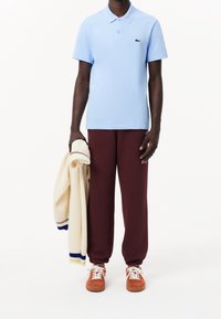 Un homme portant un polo bleu clair, un pantalon de jogging bordeaux, des baskets orange, tenant un pull en tricot crème avec des rayures bleues et jaunes.