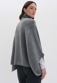 Frau, die einen grauen Wollcape mit hohem Kragen über einem weißen Langarmshirt und schwarzen Hosen trägt, und über ihre Schulter schaut.