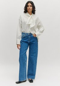 Weiße Bluse mit Nadelstreifen und Rüschekragen, kombiniert mit hoch taillierten blauen Jeans im weitem Schnitt und schwarzen Lacklederschuhen.
