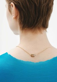 Collar de cadena de oro con un cierre rectangular descansando en la parte posterior del cuello, llevado con una blusa de tela texturizada en color verde azulado.