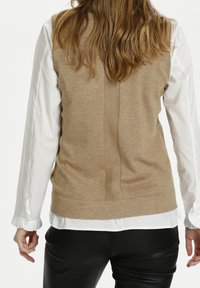 Beige pletený bezrukávový svetr s texturovaným zadním švem, přeložený přes bílou košili s volánkovými manžetami a černými koženými kalhotami.