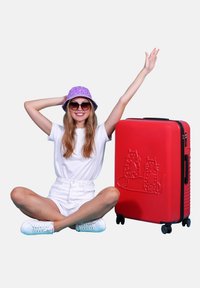 Red hard-shell suitcase with embossed cat design, featuring a retractable handle and wheels. Model in white outfit and purple bucket hat.